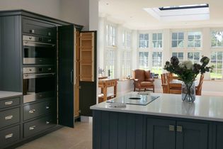 Shaker kitchen with dark grey cabinets, an open oven door, and a light-filled dining area with large windows. A vase of flowers is on the island.