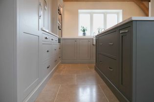 Shaker kitchen with sleek light grey cabinets around the perimeter, dark grey island. Light stone tile floor, and a white countertop. A plant by the window adds a touch of nature and warmth.