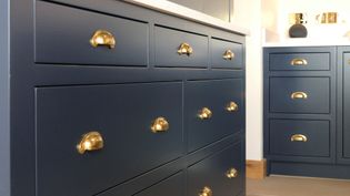 Dark blue kitchen cabinets with shiny brass cup handles create a sleek and modern look. A warm light highlights the clean lines and elegant design.