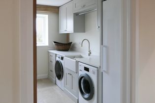 A bright utility room with two washing machines, a sink, and a wicker basket on the counter. Neutral tones and a window create an airy feel.