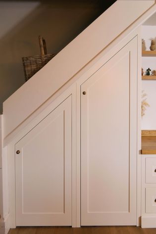 Built-in white cabinets under a staircase, featuring two angled doors with brass knobs. A wicker basket is partially visible in the corner.