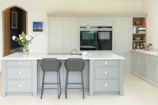 Light grey shaker style kitchen cabinetry with central island and two grey bar stools. Oven in the background. 