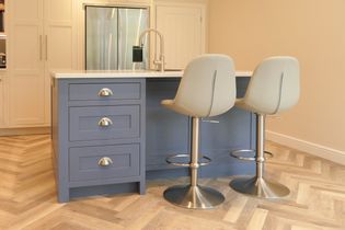 Shaker kitchen with a blue island featuring three drawers and silver handles, two beige bar stools, stainless steel appliances, and herringbone flooring.