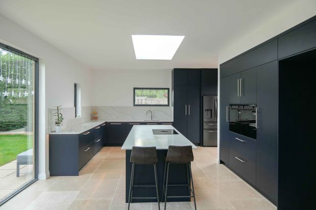 Modern dark-cabinet kitchen with a white island, two bar chairs, stainless-steel appliances, and windows overlooking a garden.