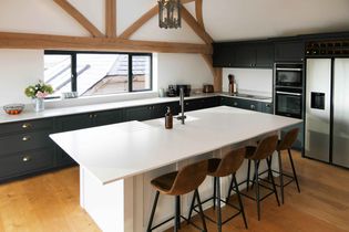 Shaker kitchen with dark cabinets, large white island and four brown barstools. Wooden beams, hanging light, and sunlit window create a cozy atmosphere.