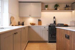 Modern kitchen with beige cabinets, wood accents, and a sleek countertop. A cutting board, fruit bowl, and knife set add warmth. Plants atop shelves.