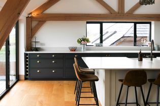 Shaker kitchen with rustic wooden beams, black cabinets, and a white island. Three bar stools add a cozy ambiance. Large windows provide natural light.