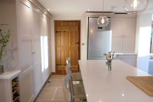 Shaker kitchen interior with neutral cabinets, wooden door, and a stainless steel fridge. A island with chairs holds a vase of flowers. Bright and elegant.