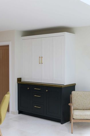 Corner of a room with a white cabinet atop a dark base with brass handles. The setting is minimalistic with a beige chair to the side.