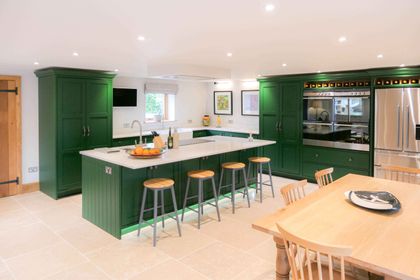A Shaker kitchen with dark green cabinets, a large central island with 4 stools, integrated appliances and a wooden table and chairs.