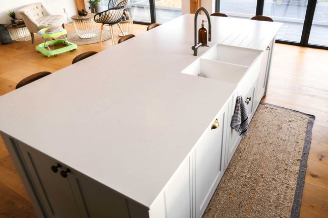 Spacious kitchen with a large white counter, built-in sink, and tap. Stylish chairs, soft-toned decor, and sunlight create a cozy ambiance.