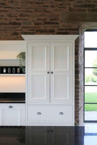 White kitchen cabinet against a rustic brick wall, with black countertop reflecting light. Nearby window offers a view of lush green lawn. Calm and modern.
