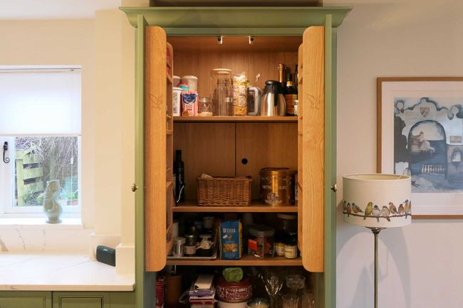 Open pantry cupboard in a shaker kitchen with jars, baskets, and food items neatly arranged. A lamp with bird illustrations and a painting nearby. Warm, organized vibe.