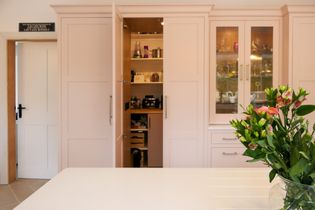 Secret pantry door open displaying different food items, a bunch of pink flowers on the worktop with the glassware cupboard behind.