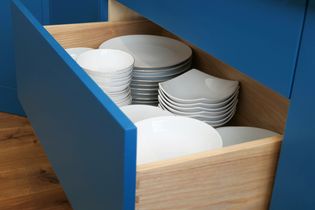 Open blue kitchen peg drawer with neatly stacked white plates and bowls on a wooden base, conveying tidiness and organization.
