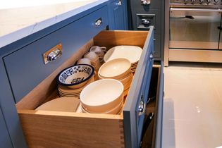 Open kitchen drawer with neatly stacked bowls, including a blue-patterned one, beside cups. Blue cabinets and an oven create a modern feel.