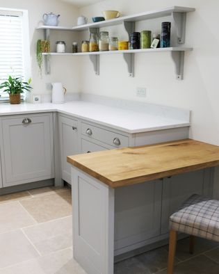 A shaker kitchen corner with light grey cabinets, a wooden countertop, open shelves holding jars and canisters, and a plaid chair. A soothing, organised space.