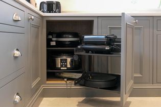 Open grey kitchen corner cabinet with pull-out shelves holding small appliances like a waffle maker and slow cooker. Organized, modern kitchen setting.