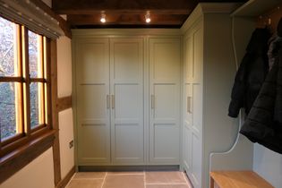 Shaker style boot room with light green built-in cabinets under warm lighting. Wooden window frames and beams add a rustic feel. Coats hang on the side.