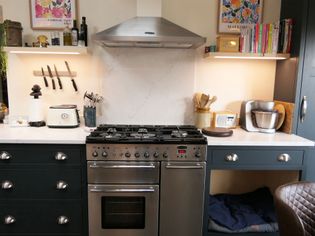 Shaker kitchen with a stainless steel gas stove, surrounded by dark cabinetry. Shelves hold cookbooks, knives, and small appliances, creating a cozy, functional space.