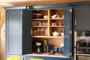 Open kitchen cabinet with blue doors reveals neatly arranged dishware, glasses, mugs, and a coffee maker on a countertop. Bright and organized atmosphere.