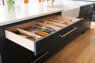 Open kitchen drawer with neatly organized cutlery and utensils, including knives and spoons, against a sleek dark cabinet and light wooden floor.