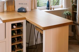 Shaker kitchen nook with a wooden counter, holding a bread box, knife set, and wine rack. Sunlit window with plants adds warmth and charm.