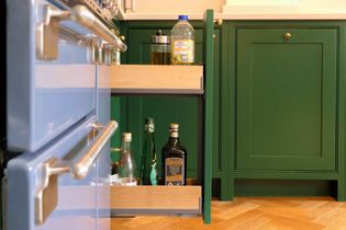 Pull-out kitchen cabinet with bottles of oil and vinegar next to a blue oven. Green shaker cabinets and wooden flooring create a vibrant atmosphere.