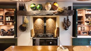 Warmly lit Shaker kitchen with a central stove, brick backsplash, open shelves, cookware, and decorative vessels.