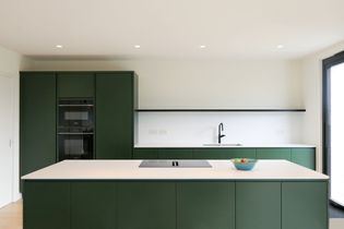 Modern kitchen with sleek green cabinetry and a minimalist design. Features a white countertop, built-in stovetop, black faucet, and a bowl of fruit.