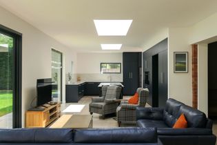 Modern open-plan living space with sleek kitchen, skylights, and a garden view. Blue sofas with orange cushions and plaid chairs add warmth.