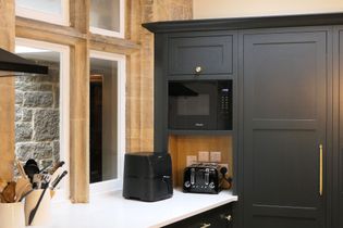 Shaker kitchen with dark cabinetry, built-in microwave, toaster, and air fryer on a white countertop. Stone wall and windows add rustic charm.