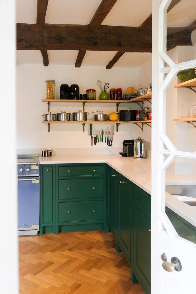 Shaker kitchen with green cabinets, wooden shelving, and a blue stove. Rustic beams, white counters, pots, and colorful jars create a warm, inviting atmosphere.