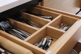 Open wooden drawer with compartments holding spoons, forks, knives, small cutlery, and various kitchen tools.