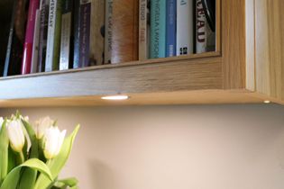 A wooden bookshelf holds various cookbooks with a warm under-shelf light illuminating a bouquet of white tulips below, creating a cozy ambiance.