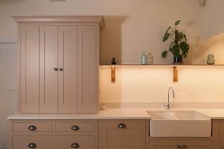 Light beige kitchen with a white countertop and farmhouse sink. Cabinets have simple knobs, and a shelf holds green plants and small bottles, creating a calm, warm tone.