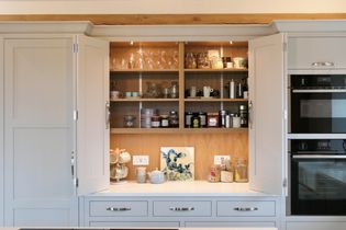 Open light grey countertop cupboard with bi fold doors revealing shelves stocked with glassware, pottery, and jars. Counters display mugs and a colorful art piece.