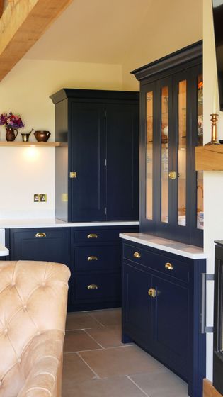 A cozy kitchen corner featuring navy blue cabinets with brass handles, a tan tufted armchair, and a warm beige wall adorned with small decorative items.