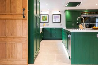 Shaker kitchen with vibrant green cabinetry, a wooden door on the left, light stone floor tiles, and framed artwork on white walls, creating a stylish and inviting atmosphere.