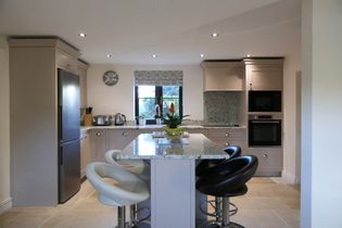 Shaker kitchen with an island, surrounded by chairs. Neutral-toned cabinets, stainless steel appliances, and a plant add a cozy touch. Bright lighting with a window view.