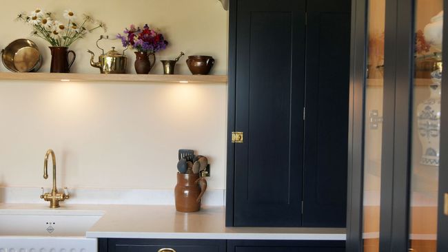 A cosy kitchen features a dark cabinet, a wooden shelf with brass teapots and fresh flowers. Soft lighting and a brass faucet add warmth.