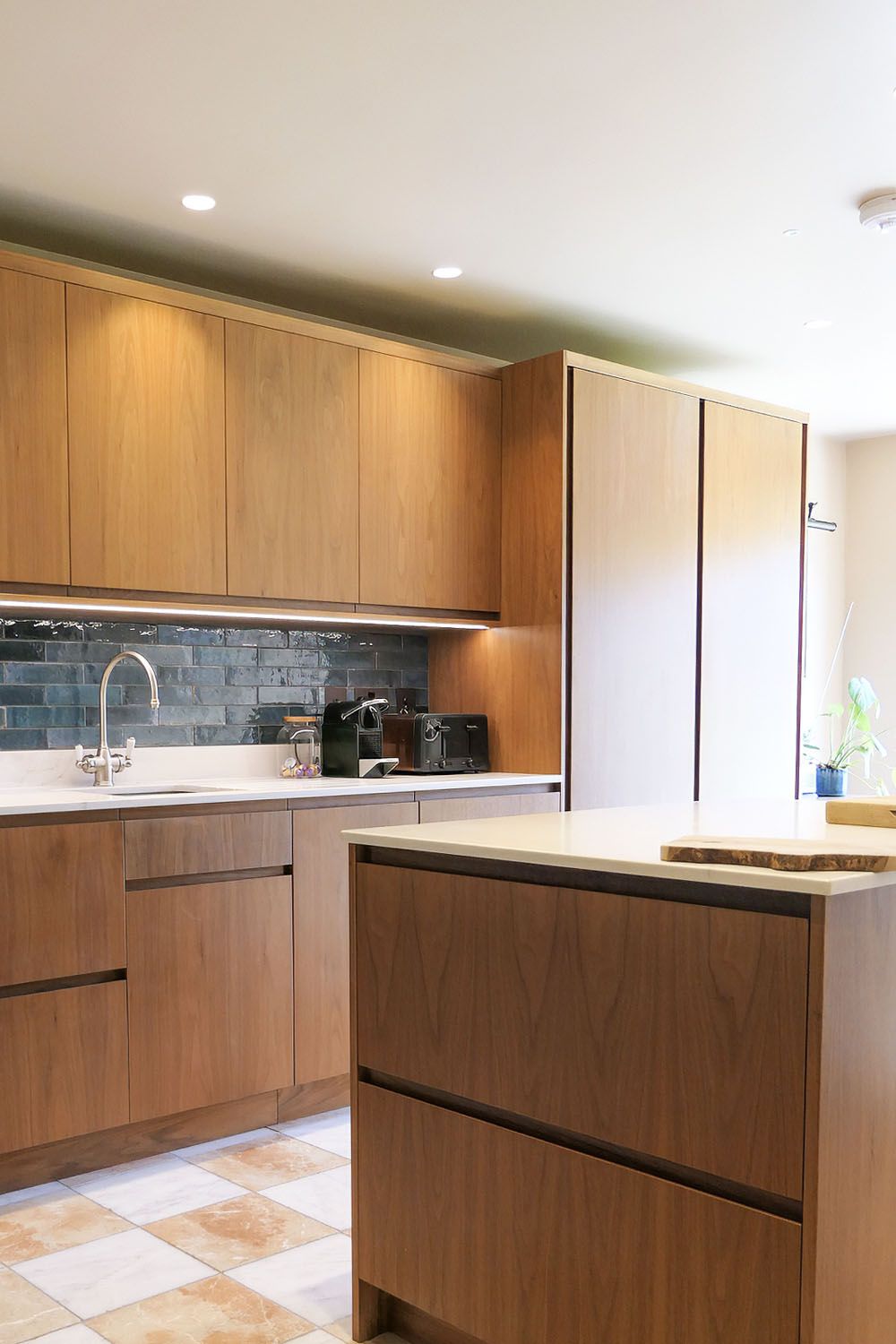 Modern walnut kitchen with a white island, dark tiled backsplash, sink, coffee machines, and a potted plant.