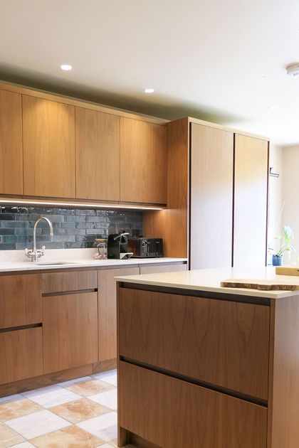 Modern walnut kitchen with a white island, dark tiled backsplash, sink, coffee machines, and a potted plant.