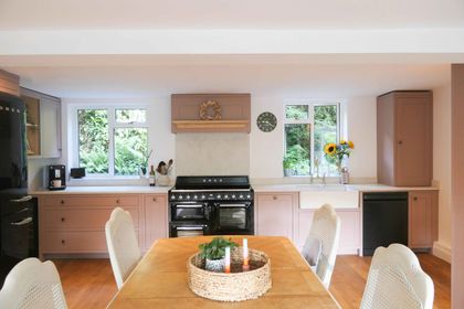 Blush pink Shaker kitchen with black appliances, two kitchen windows with a vase of sunflowers on the white worktop. A wooden dining table is in the foreground.