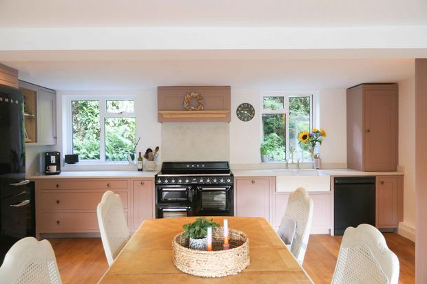 Blush pink Shaker kitchen with black appliances, two kitchen windows with a vase of sunflowers on the white worktop. A wooden dining table is in the foreground.