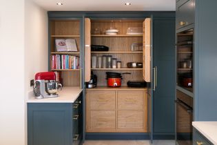 Shaker kitchen with blue cabinets and open wooden pantry. Shelves hold cookbooks, jars, and cookware. Red mixer on countertop adds a vibrant touch.