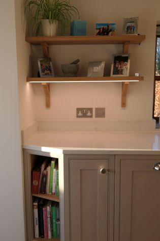 Wooden shelves above a beige kitchen counter display potted plants and framed photos. Below, books fill a corner shelf, creating an inviting ambiance.