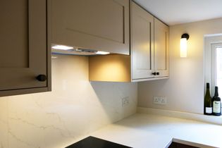 A Shaker kitchen with light-coloured cabinets, a white countertop, and under-cabinet lighting. There are wine bottles on the counter near a window.