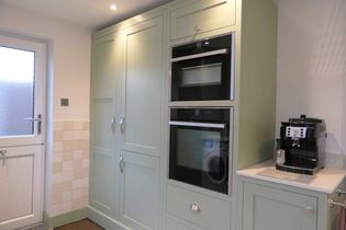 Shaker kitchen with soft green cabinets featuring built-in ovens. A silver coffee machine sits on the countertop. Natural light from a frosted glass door.