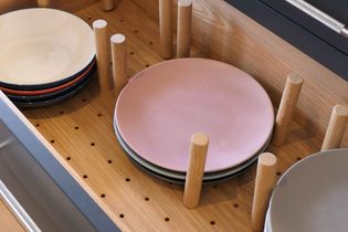 Wooden drawer with peg board design, holding stacks of pastel pink and neutral plates separated by wooden dowels. Neat, organized, minimalist tone.